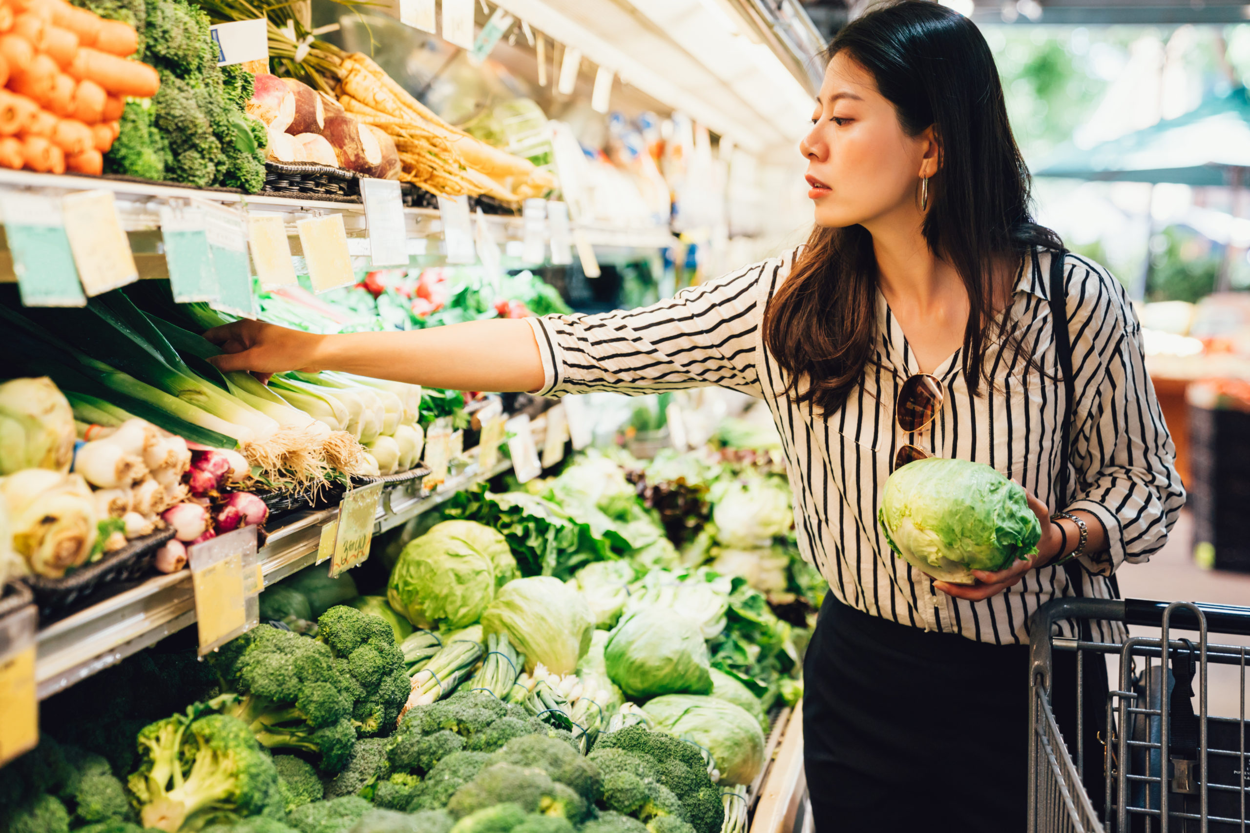 Asian American woman holding cabbage in one hand and picking spring onions with the other hand in produce section