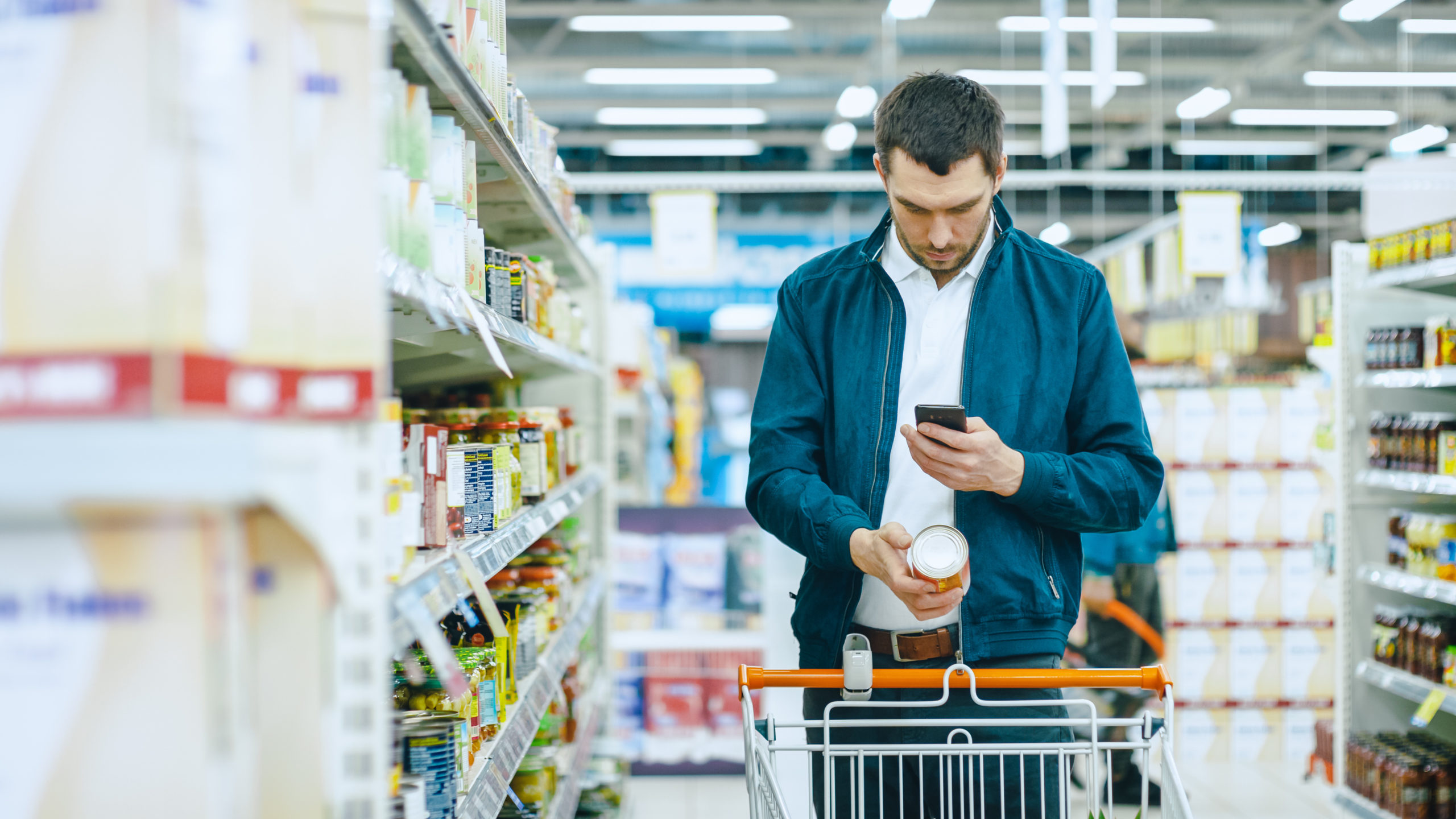At the Supermarket: Handsome Man Uses Smartphone and Takes Picture of the Can of Goods. He's Standing with Shopping Cart in Canned Goods Section.