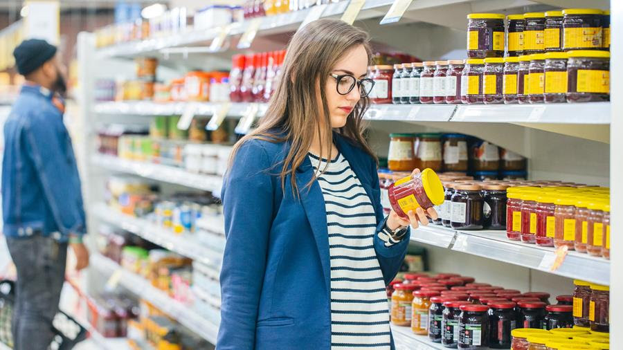Caucasian woman with straight hair is wearing a blue blazer, striped shirt is seen holding a jar of honey standing in a supermarket