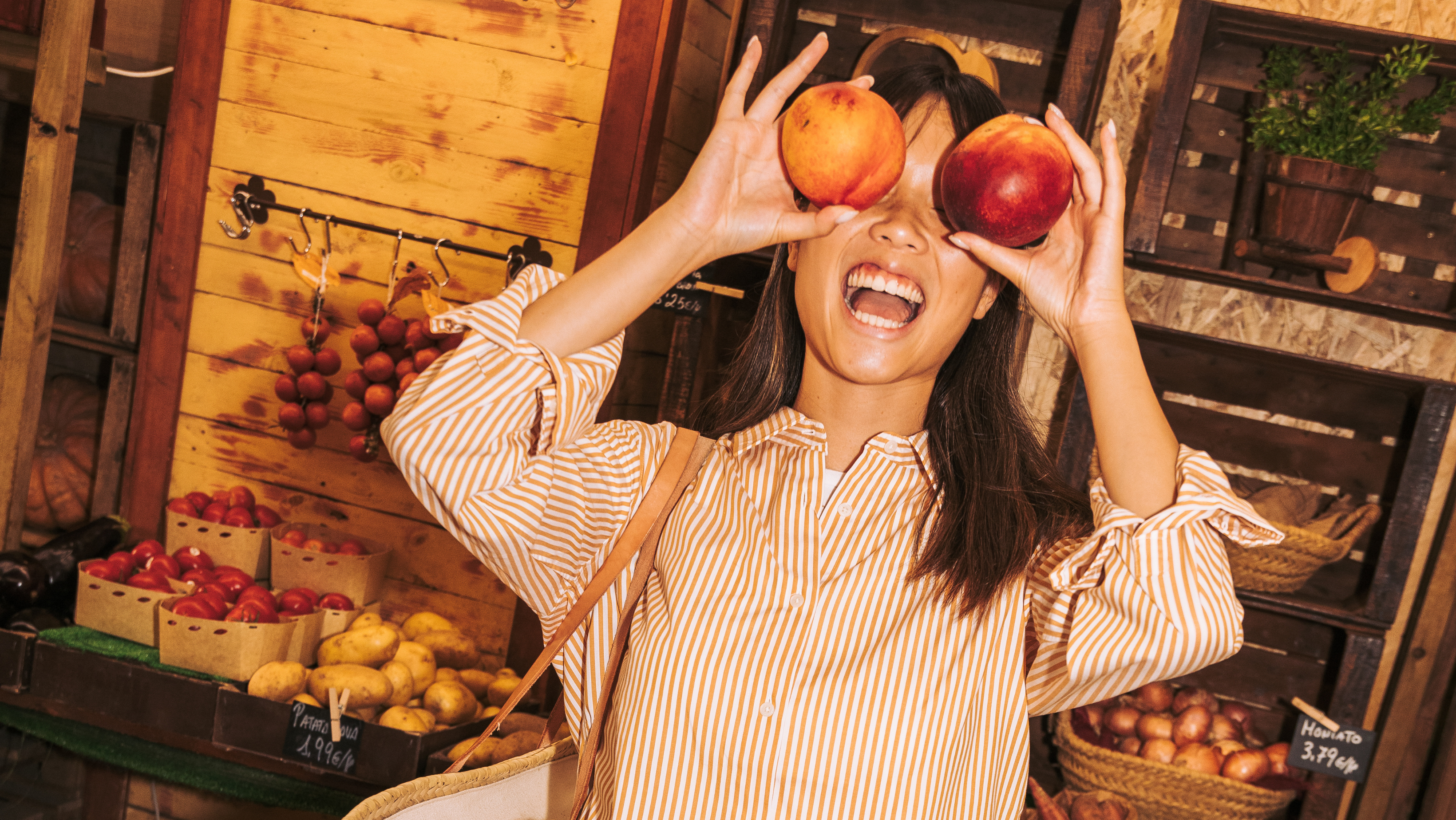 A young woman at a produce market laughs and holds two nectarines in front of her eyes as a joke