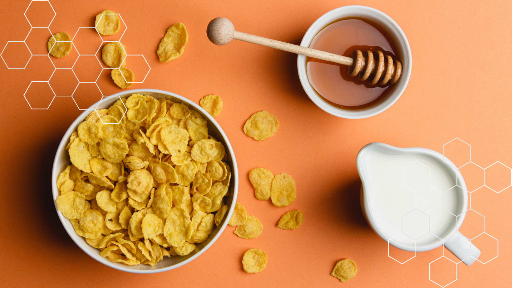 Looking down on an orange table top, into a bowl of flaked corn cereal, a small pitcher of cold white milk and a bowl of delicious honey. There decorations of honeycomb shapes overlaid in white on the upper left and lower right corners of the image.
